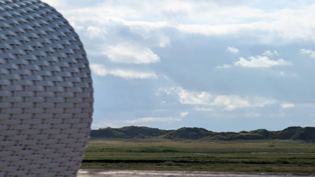 Close-up of the woven white side panel of a beach chair with a wide view of green salt marshes, gentle dunes and a cloudy sky in the background.