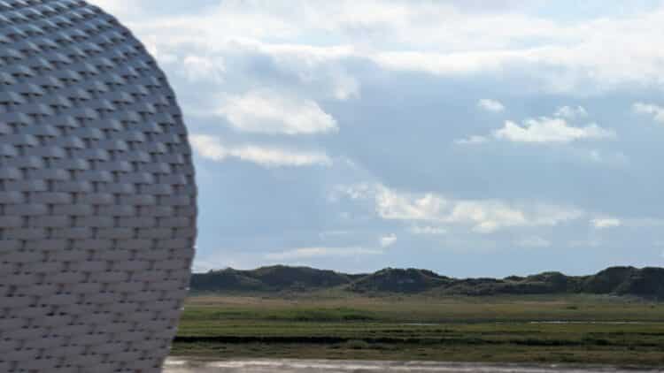 Nahaufnahme des geflochtenen weißen Seitenteils eines Strandkorbs mit weitem Ausblick auf grüne Salzwiesen, sanfte Dünen und bewölkten Himmel im Hintergrund.